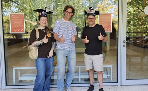 Auf dem Bild sieht man von links nach rechts, Melinda Braun, Matthias W&ouml;lfel und Thorsten Zyloswki. Melinda Braun und Thorsten Zylowski tragen beide ihre Doktorh&uuml;te. Alle drei zeigen ihre Daumen nach oben. Das Bild wurde vor einer Glasfront auf einem Balkon der HKA aufgenommen. 