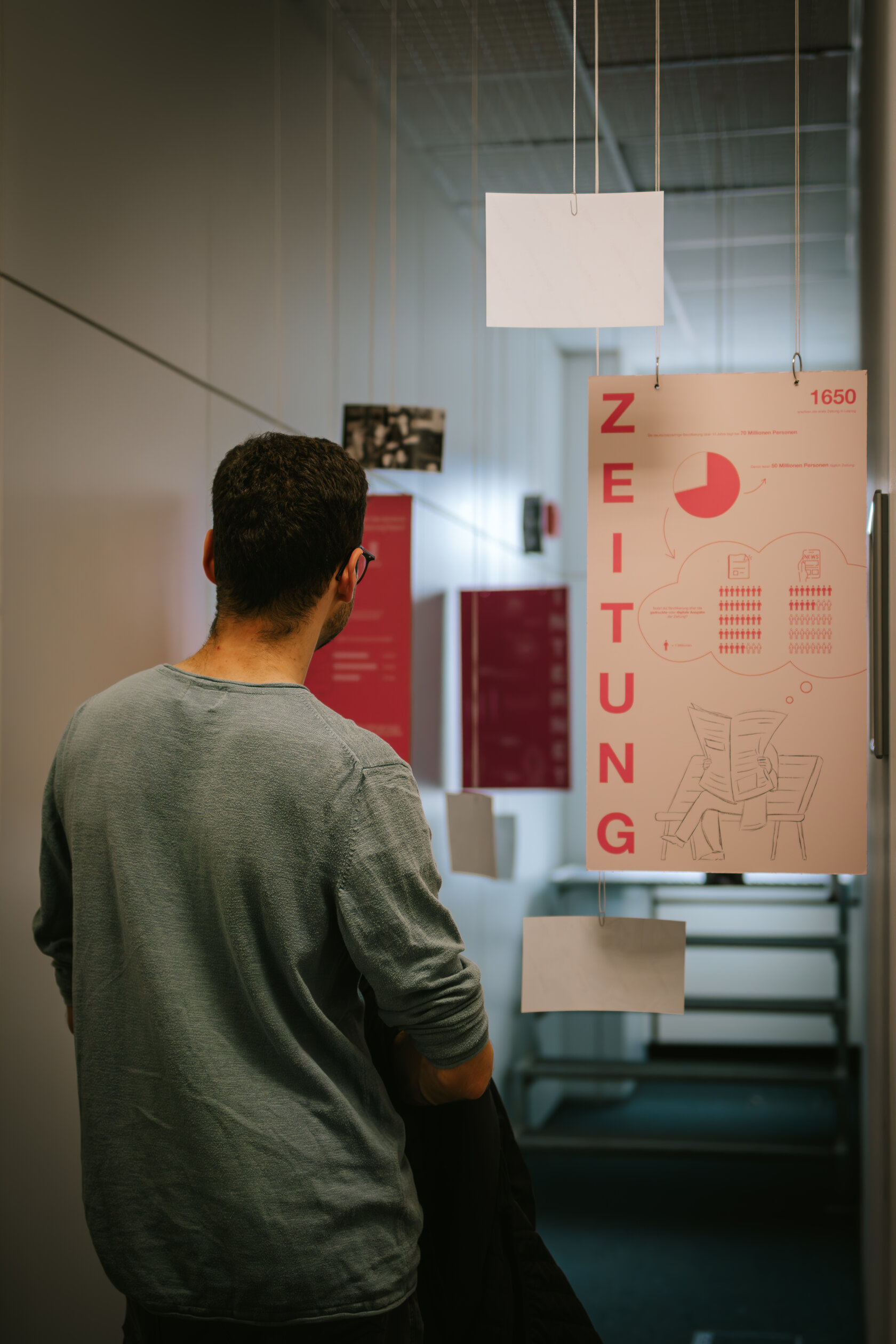 Mann steht vor Ausstellungsstück „Die Entwicklung der öffentlichen Medien“ von Samira Weigand. Mehrere Plakete hängen in einem schmalen Gang von der Decke.