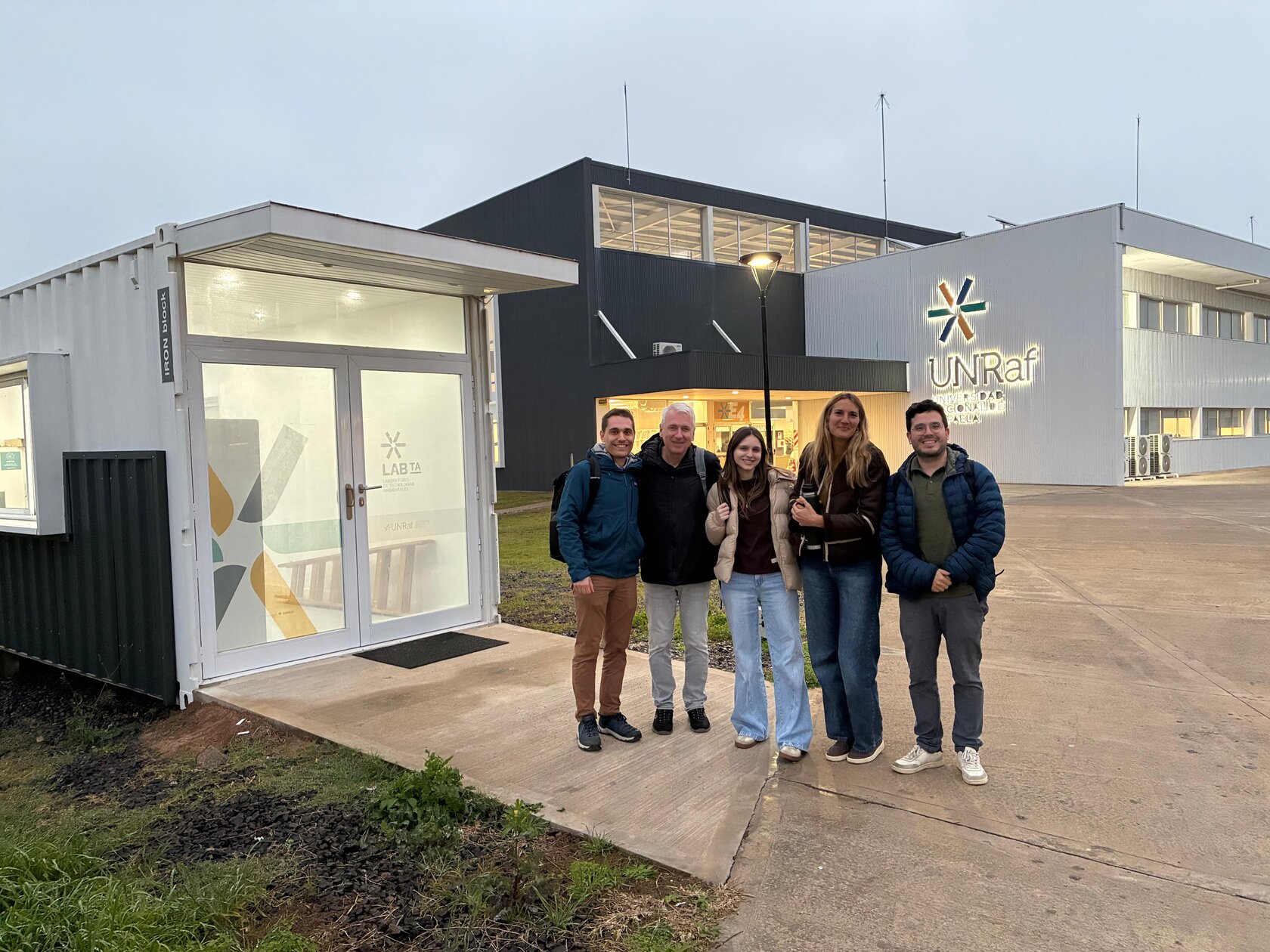 Fünf Personen stehen lächelnd vor einem Laborcontainer mit der Aufschrift "LAB TA" auf dem Campus der UNRaf, im Hintergrund das Hauptgebäude mit UNRaf-Logo.