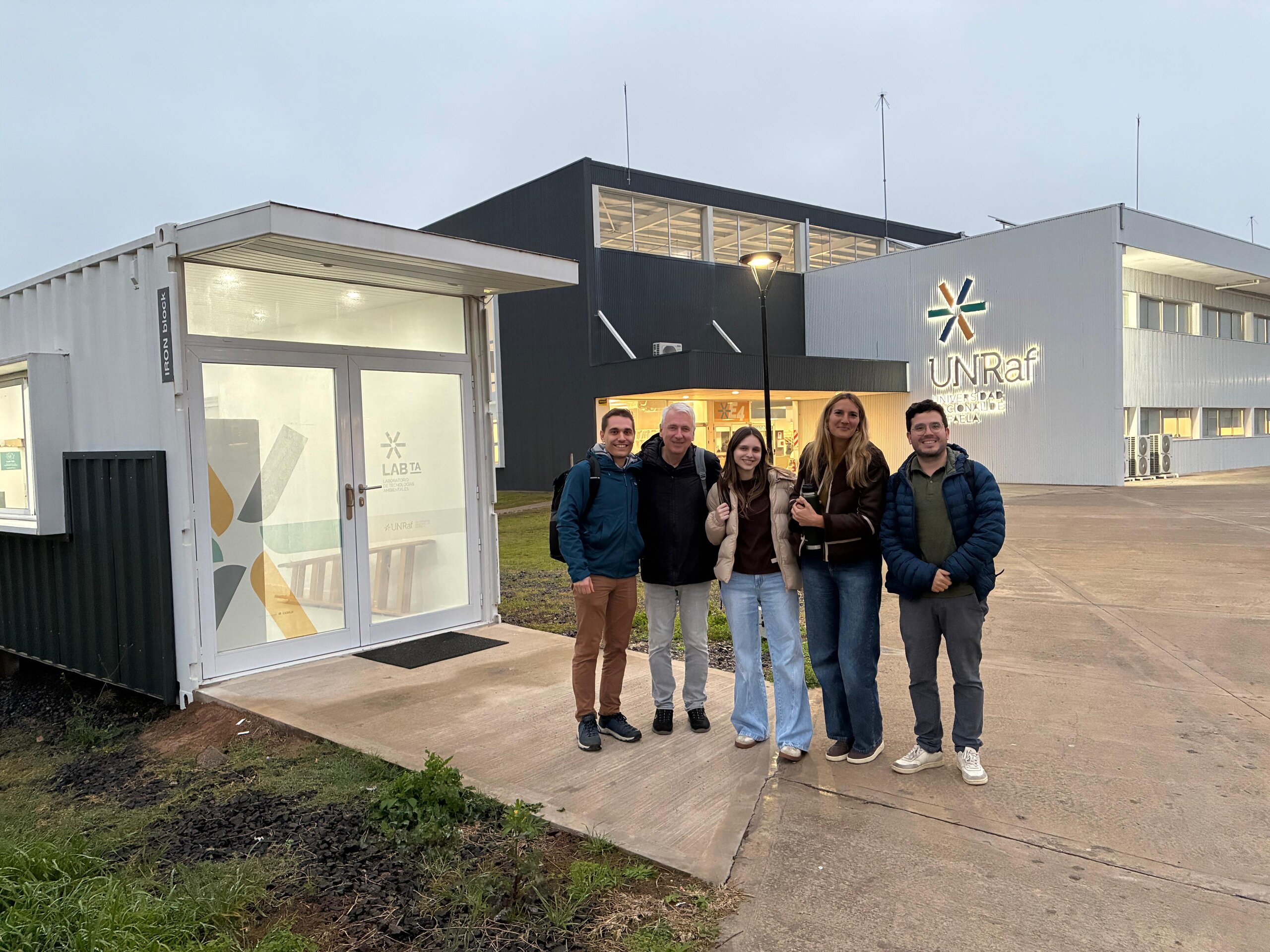Fünf Personen stehen lächelnd vor einem Laborcontainer mit der Aufschrift "LAB TA" auf dem Campus der UNRaf, im Hintergrund das Hauptgebäude mit UNRaf-Logo.
