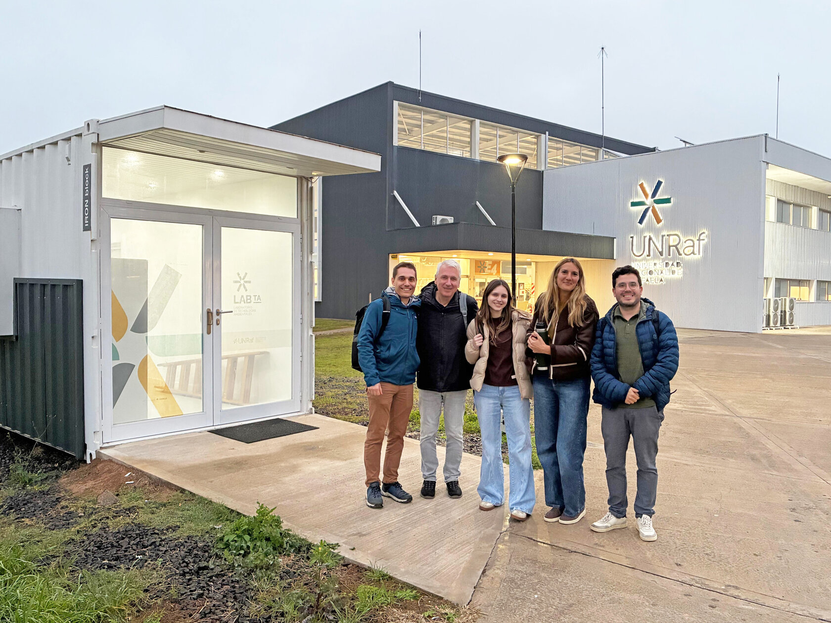 Fünf Personen stehen lächelnd vor einem Laborcontainer mit der Aufschrift "LAB TA" auf dem Campus der UNRaf, im Hintergrund das Hauptgebäude mit UNRaf-Logo.