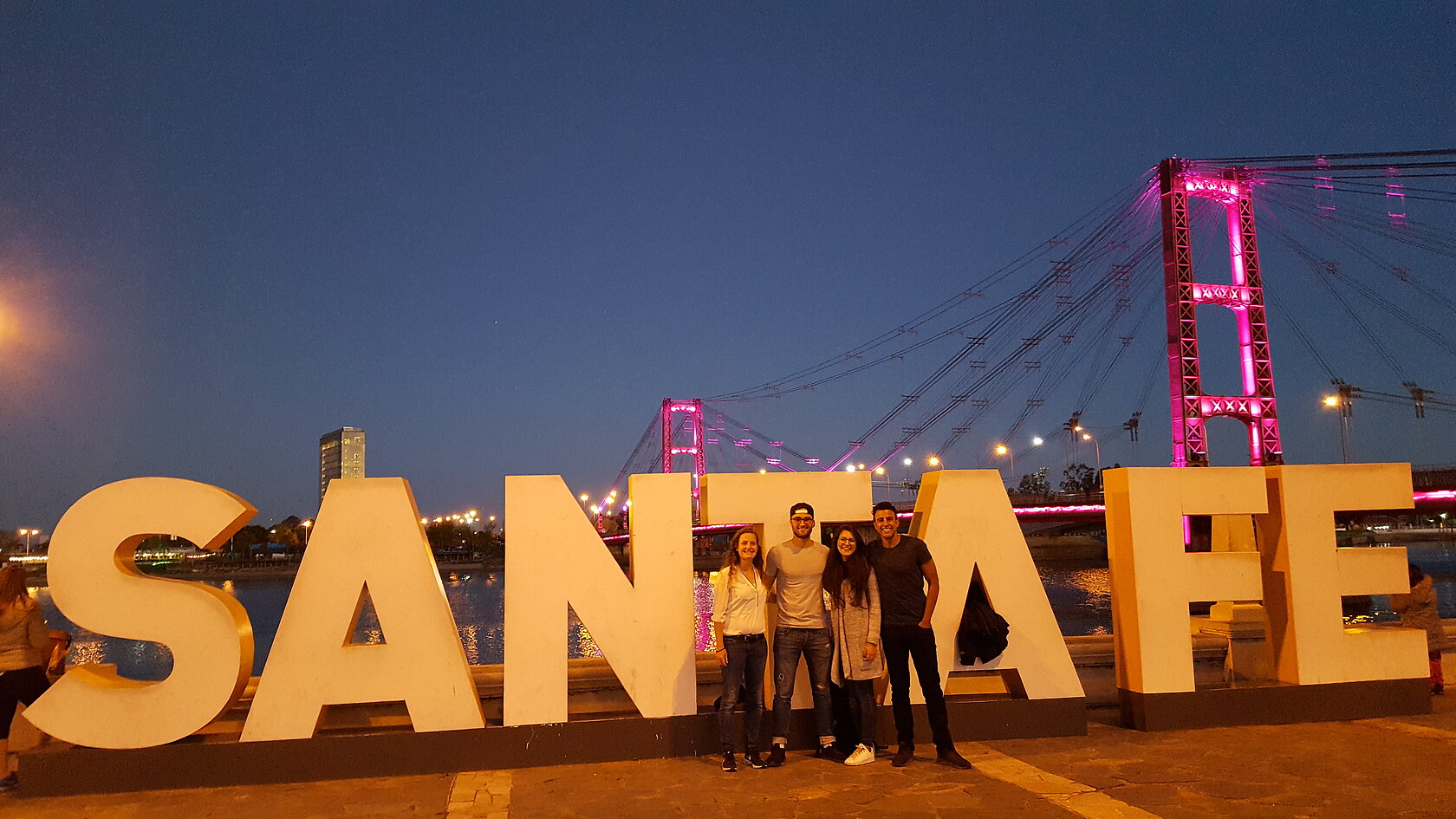 students stand in front of a bridge, at a sculpture of large letters forming the word "Santa Fe"