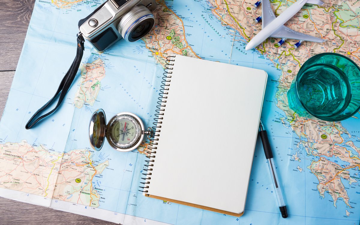Ein leeres Notizbuch liegt auf einer Landkarte. Daneben befinden sich ein Stiff, ein Kompass, ein Fotoapparat, ein Flugzeugmodell und ein Glas Wasser.