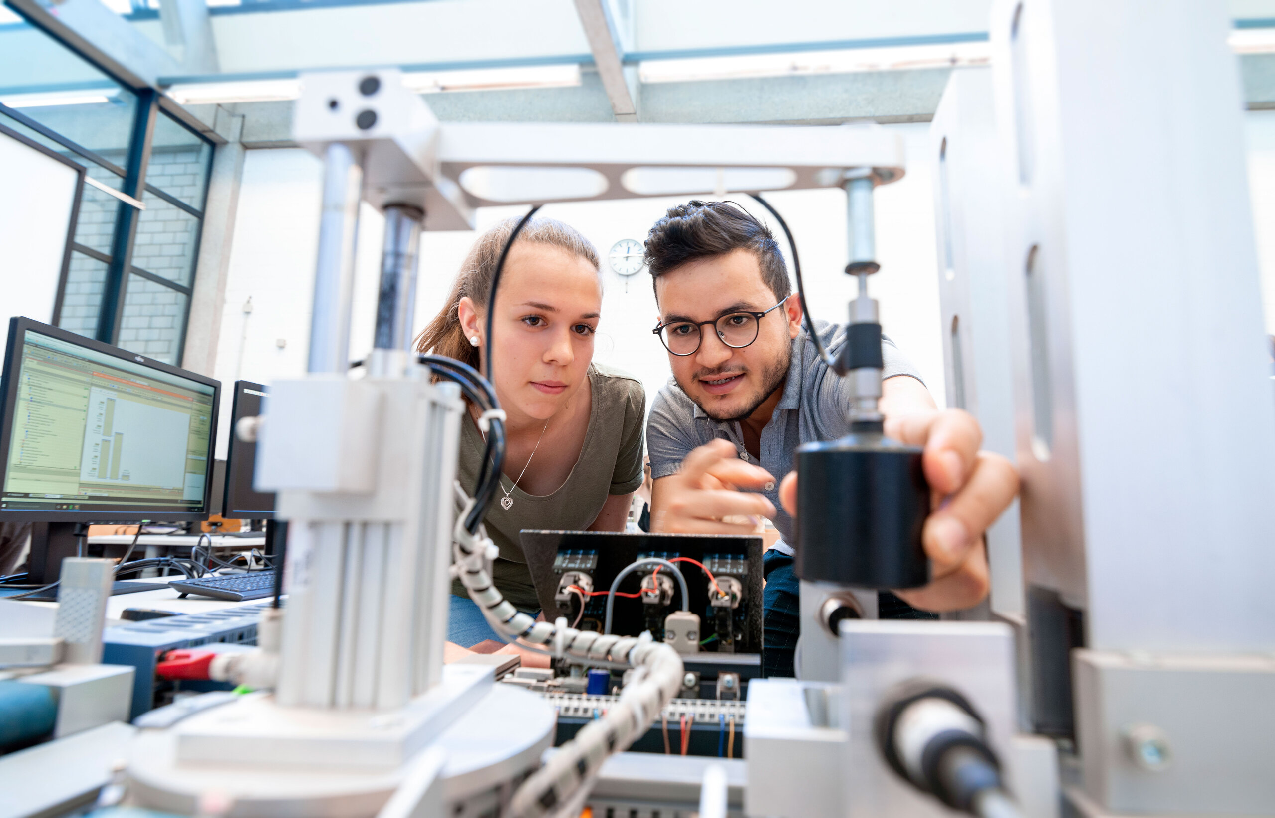 two students working on a machine at the lab, with computer in the background