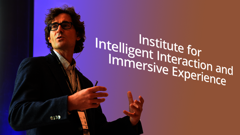 Das Bild zeigt Matthias W&ouml;lfel bei einem Vortrag vor einer Leinwand, auf der "Institute for Intelligent Interaction and Immersive Experience" geschrieben ist.