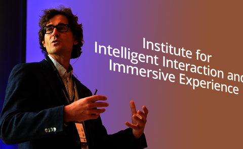 Das Bild zeigt Matthias Wölfel bei einem Vortrag vor einer Leinwand, auf der "Institute for Intelligent Interaction and Immersive Experience" geschrieben ist.