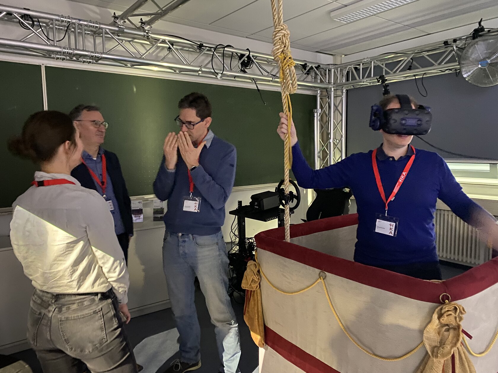 A person stands in a replica balloon basket and wears virtual reality goggles to experience a VR application. Several visitors stand next to it and watch the demonstration in the iXperience Lab at Karlsruhe University of Applied Sciences.