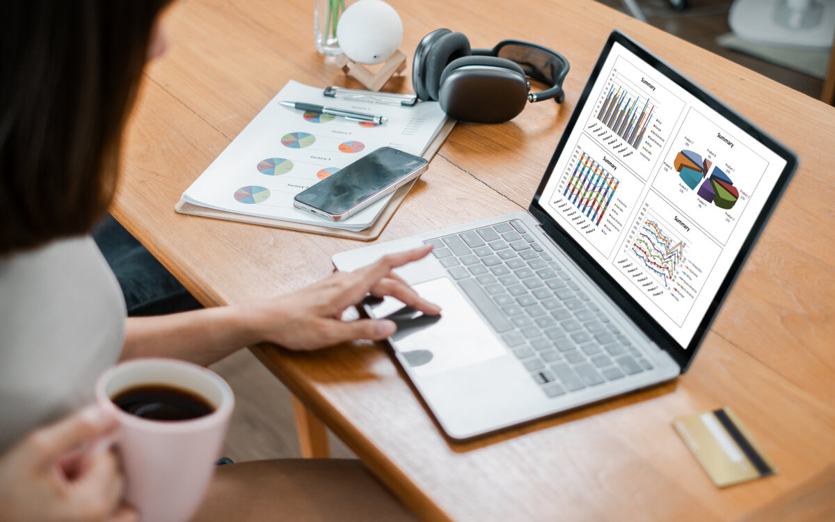 Businesswoman working on financial data analysis with a laptop, coffee, and documents on a wooden desk in a modern office setting.