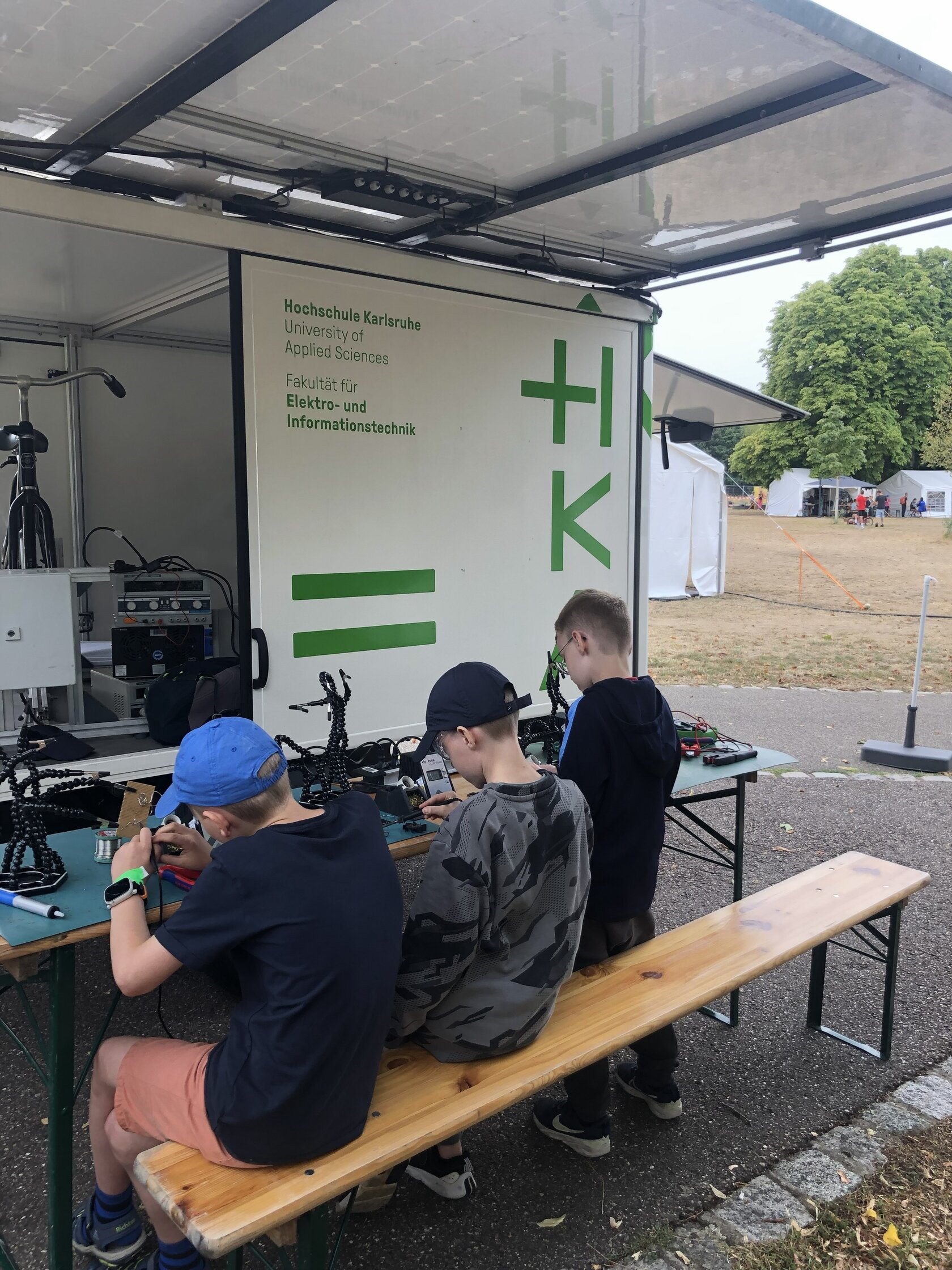 Drei Kinder sitzen auf einer Bank vor dem HKA-Ausstellungsfahrzeug und löten elektronische Bauteile.