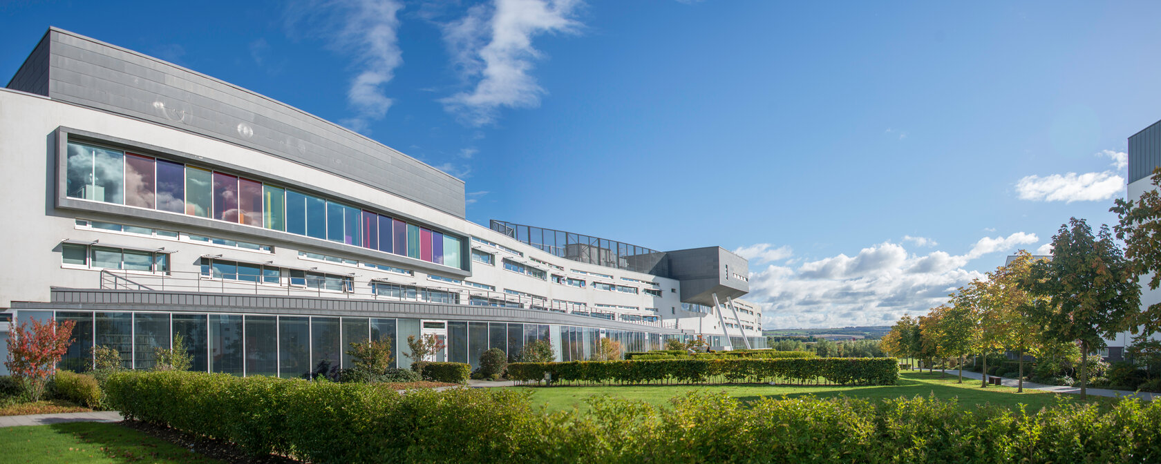 modern building with glass face, in front of this green campus with trees and hedges 