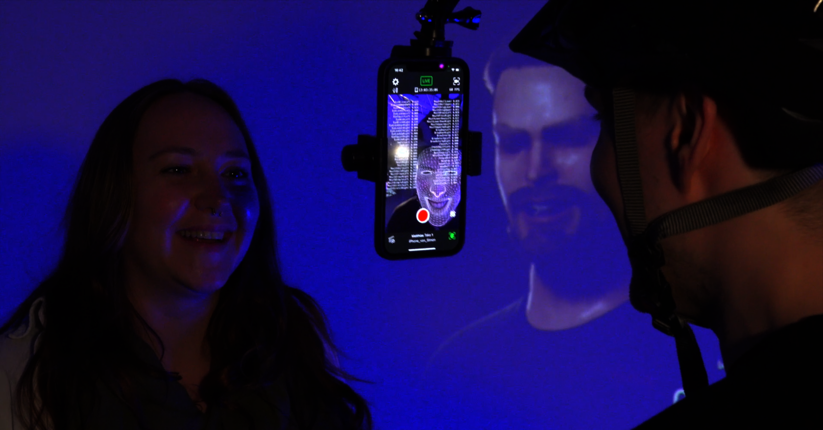 Das Bild zeigt eine junge Frau, die lächelnd in eine Smartphone-Kamera blickt. Auf dem Smartphone wird ein Gittermodell ihres Gesichts angezeigt, das in Echtzeit analysiert wird. Im Hintergrund ist ein digitaler Avatar auf einem Monitor zu sehen, der die Gesichtszüge der Frau nachahmt.