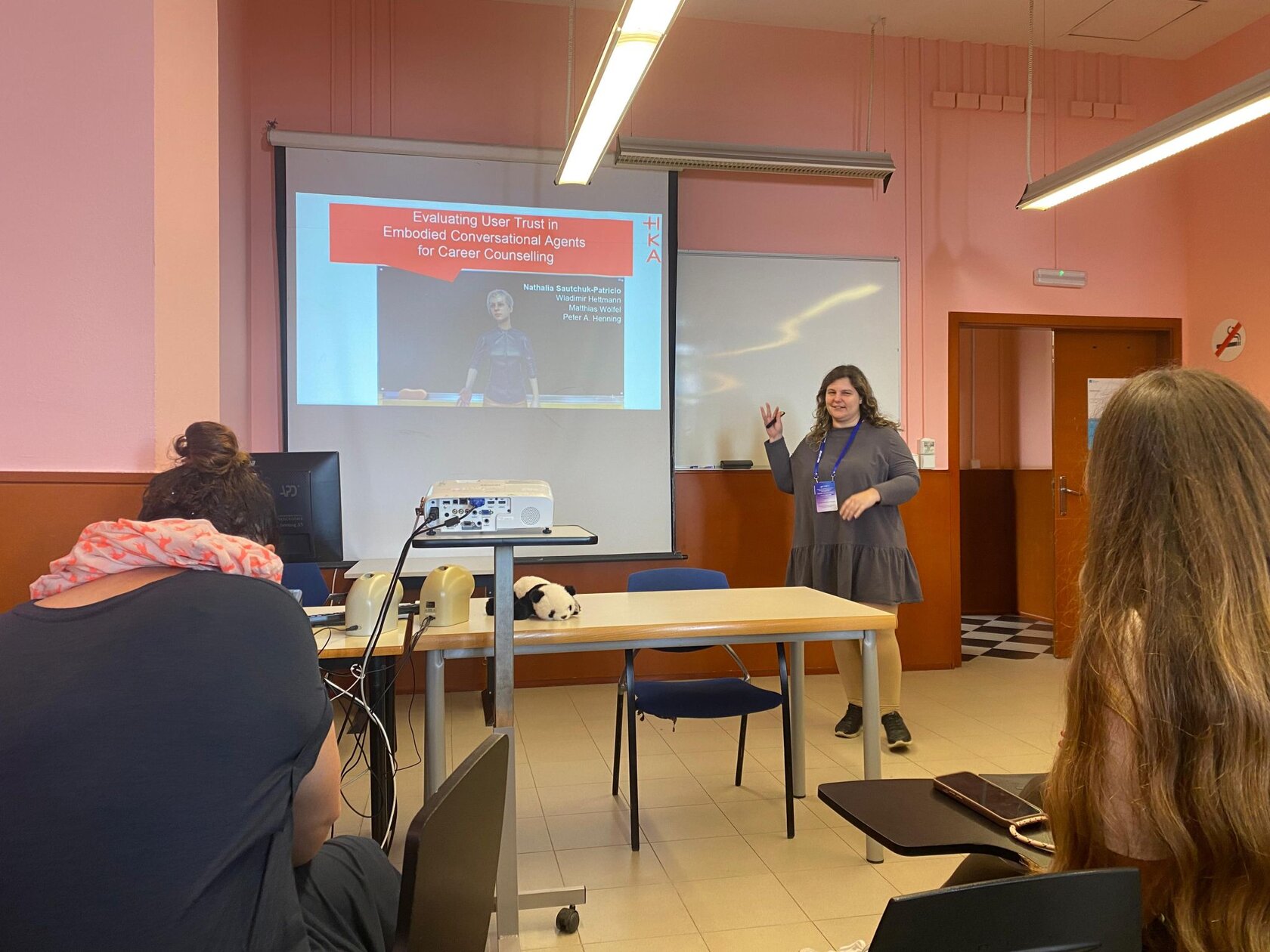 Nathalia Sautchuk-Patricio speaks in front of a presentation slide showing her research on building trust in AI-based career assistants. She turns to the audience and gestures.