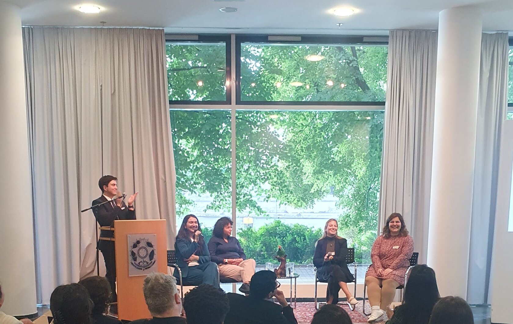 Vier Teilnehmerinnen sitzen in einer Diskussionsrunde, ein Teilnehmer steht hinter einem Redepult und klatscht.