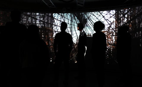 The image shows a group of people in a dark 180-degree projection environment. They are looking at a large-scale projection showing the structure of a historic airship. The scene is kept in muted colors, the people are visible as silhouettes, while the projection is the main focus.