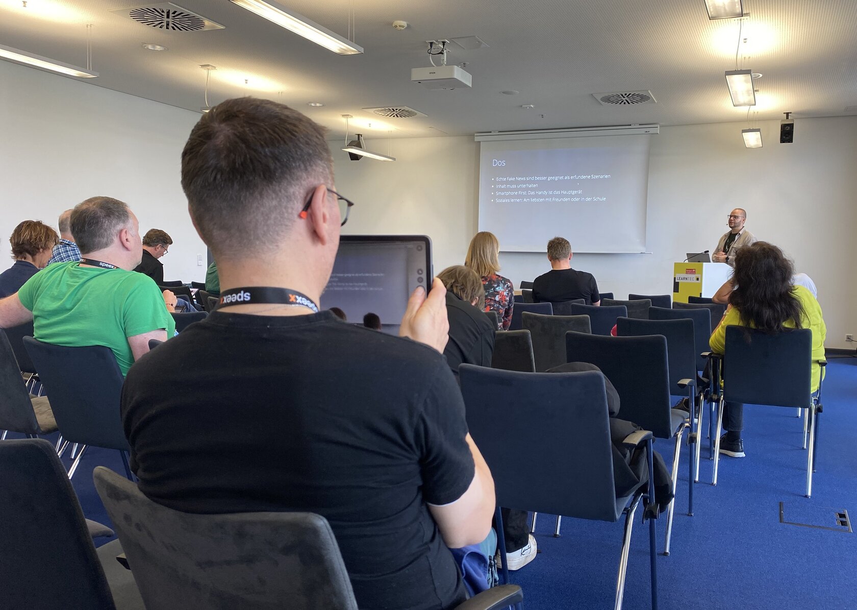 Teilnehmende der LEARNTEC 2026 verfolgen eine Präsentation im Kongressprogramm. Im Vordergrund sitzt ein Besucher mit einem Tablet, auf dem Inhalte des Vortrags angezeigt werden. Im Hintergrund sind weitere Zuhörerinnen und Zuhörer sowie Niklas Oesterle vor einer Projektionsfläche zu sehen.