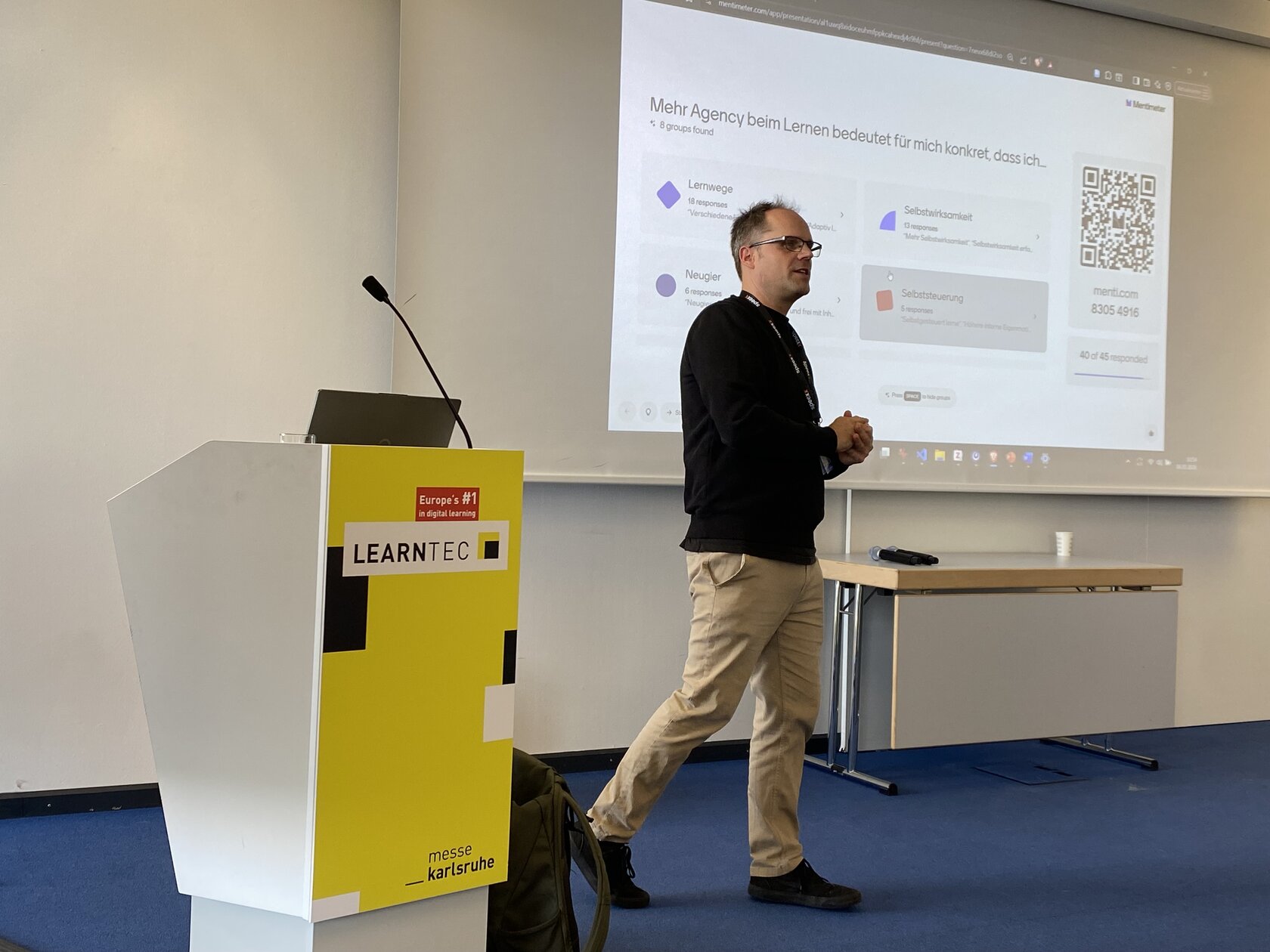 Thorsten Zylowski steht neben einem Rednerpult mit der Aufschrift LEARNTEC 2026. Hinter ihm ist eine Leinwand, auf der seine Präsentationsfolien zu sehen sind. Er blickt aufmerksam in die Menge.