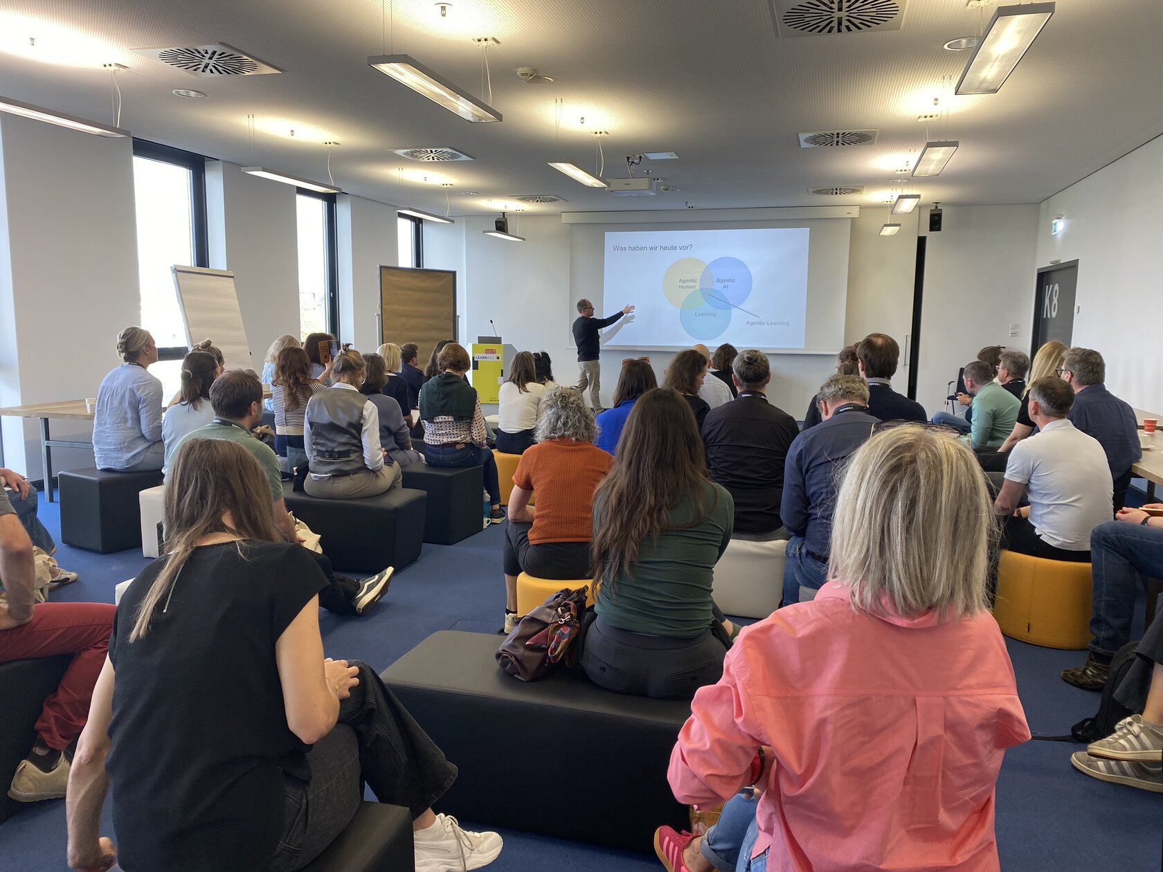 IIIX employee Thorsten Zylowski stands in front of a large group of people sitting on chairs. He points with his hand to a screen with his workshop slides.