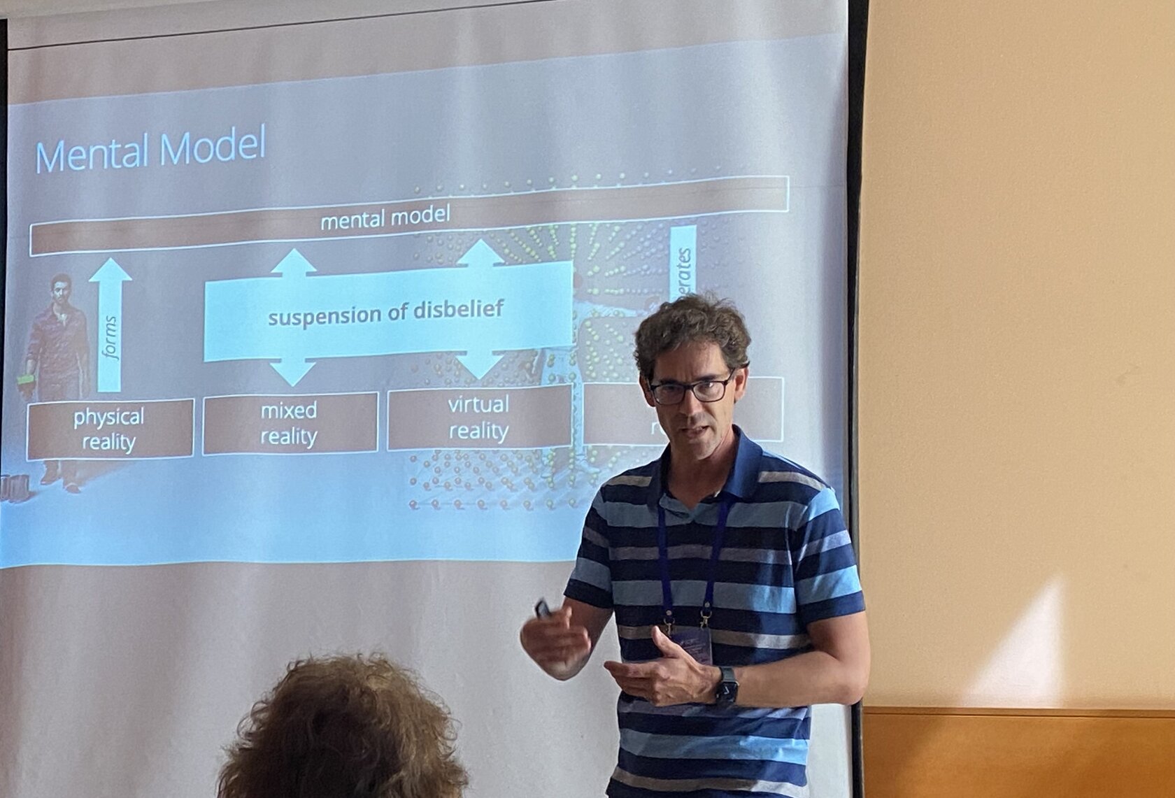 Prof. Dr. Matthias Wölfel stands in front of a screen in a lecture room and speaks into the microphone. A presentation slide from his invited speech on virtual reality and the VR Campus can be seen in the background.