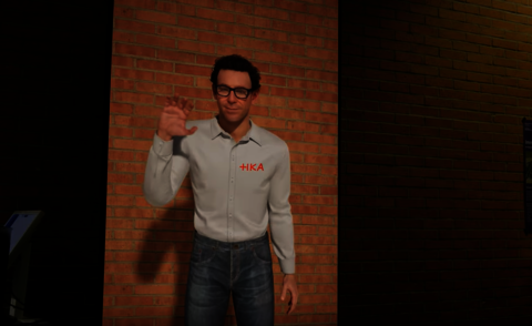 The picture shows the Embodied Conversational Agent of the IIIX, depicted as a life-size virtual character with short brown hair, glasses, a white shirt with the HKA logo and blue jeans. The agent stands in front of a brick wall and raises his hand in a friendly greeting.
