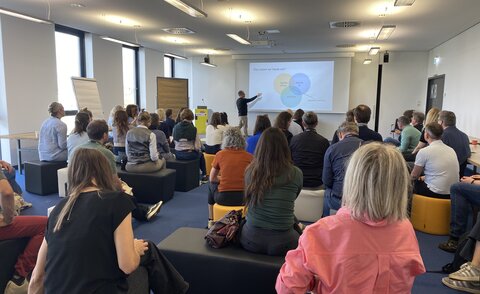 IIIX employee Thorsten Zylowski stands in front of a large group of people sitting on chairs. He points with his hand to a screen with his workshop slides.