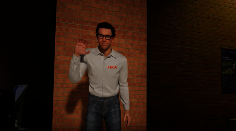 Auf dem Bild ist der Embodied Conversational Agent des IIIX zu sehen, dargestellt als lebensgro&szlig;er virtueller Charakter mit kurzen braunen Haaren, Brille, wei&szlig;em Hemd mit HKA-Logo und blauer Jeans. Der Agent steht vor einer Backsteinwand und hebt freundlich die Hand zum Gru&szlig;.